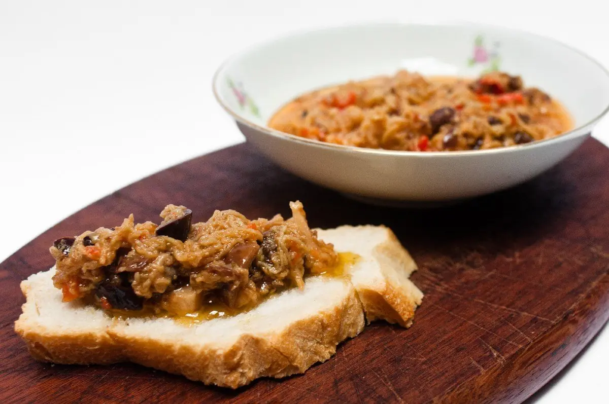 Caponata numa tigela e servida sobre fatias de pão italiano, na tábua de madeira