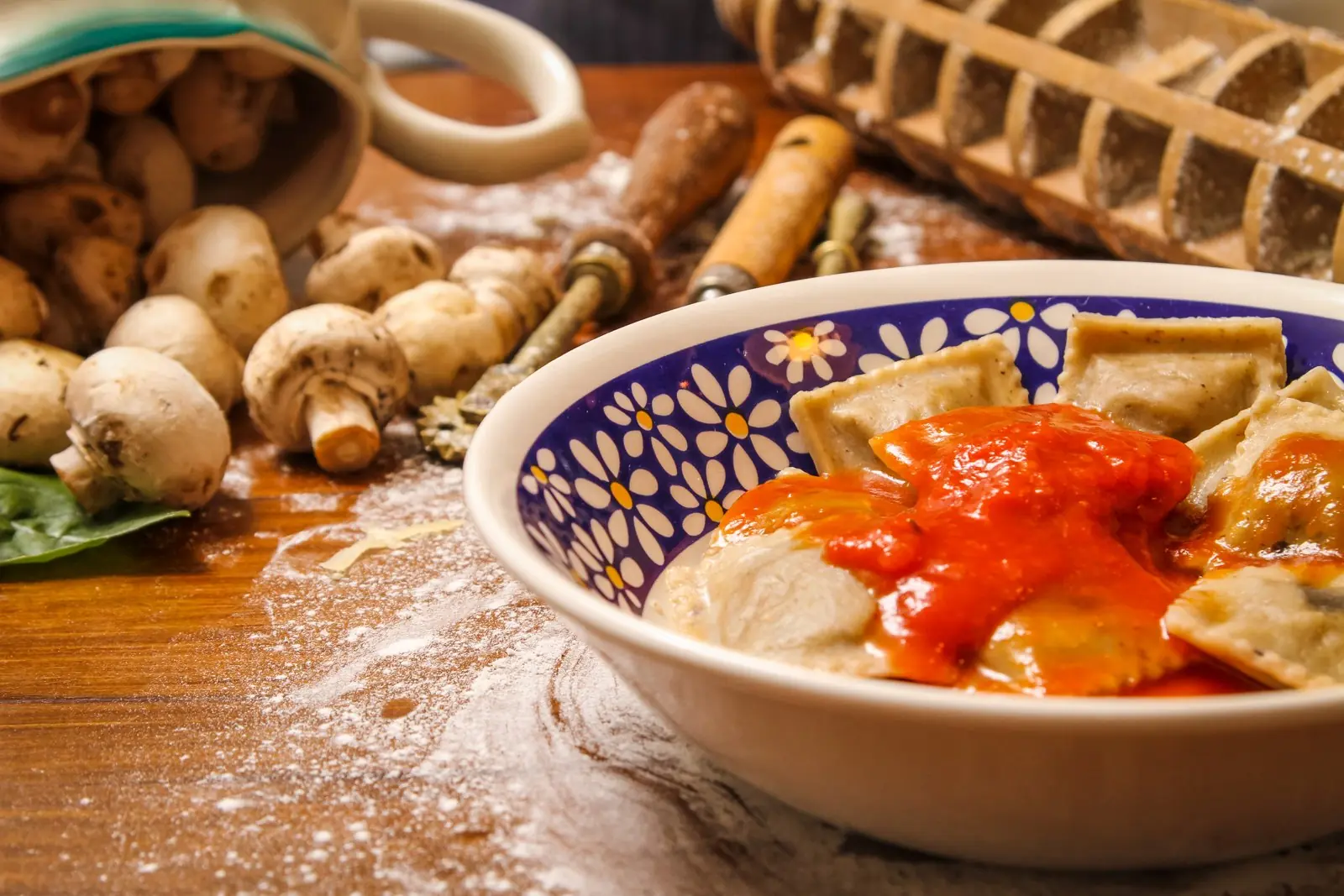 Ravióli ao pomodoro numa tigela de cerâmica pintada, com cogumelos e farinha na bancada