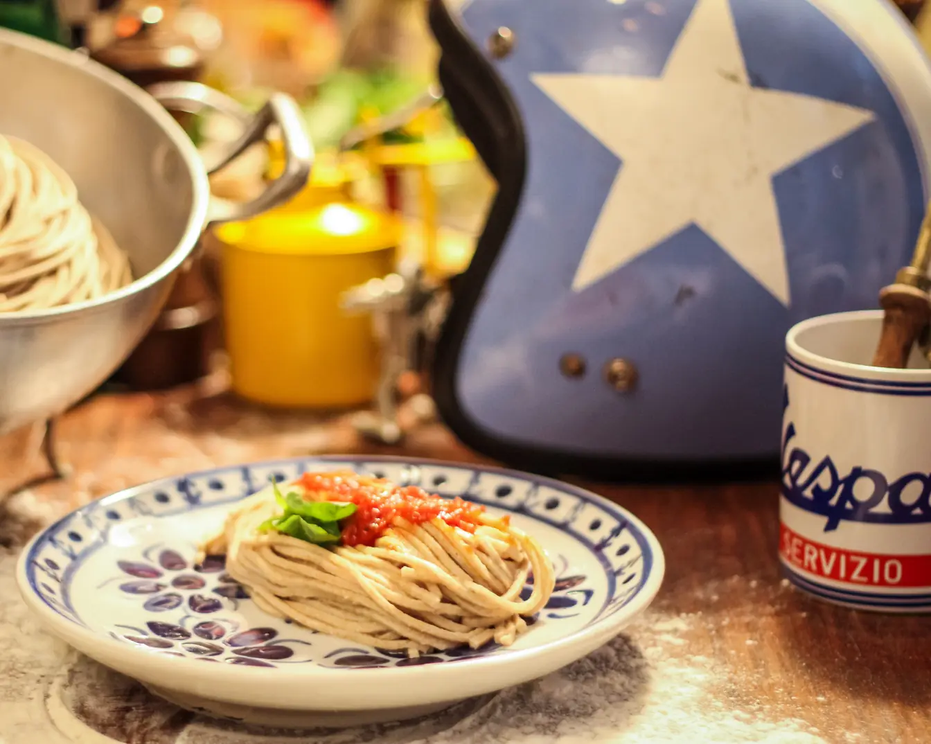 Spaghetti fresco ao pomodoro em prato de cerâmica azul e branca, com um capacete e uma caneca Vespa ao fundo, na bancada do Primo