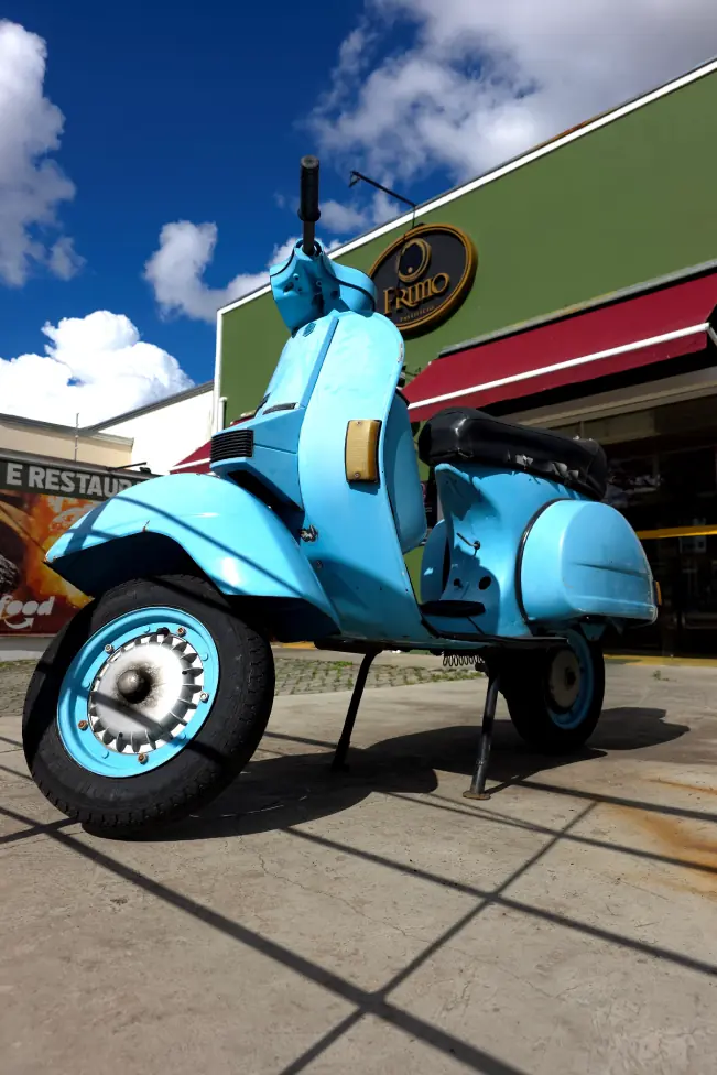 Vespa azul estacionada em frente à loja do Primo em Sorocaba, com o letreiro oval na fachada verde